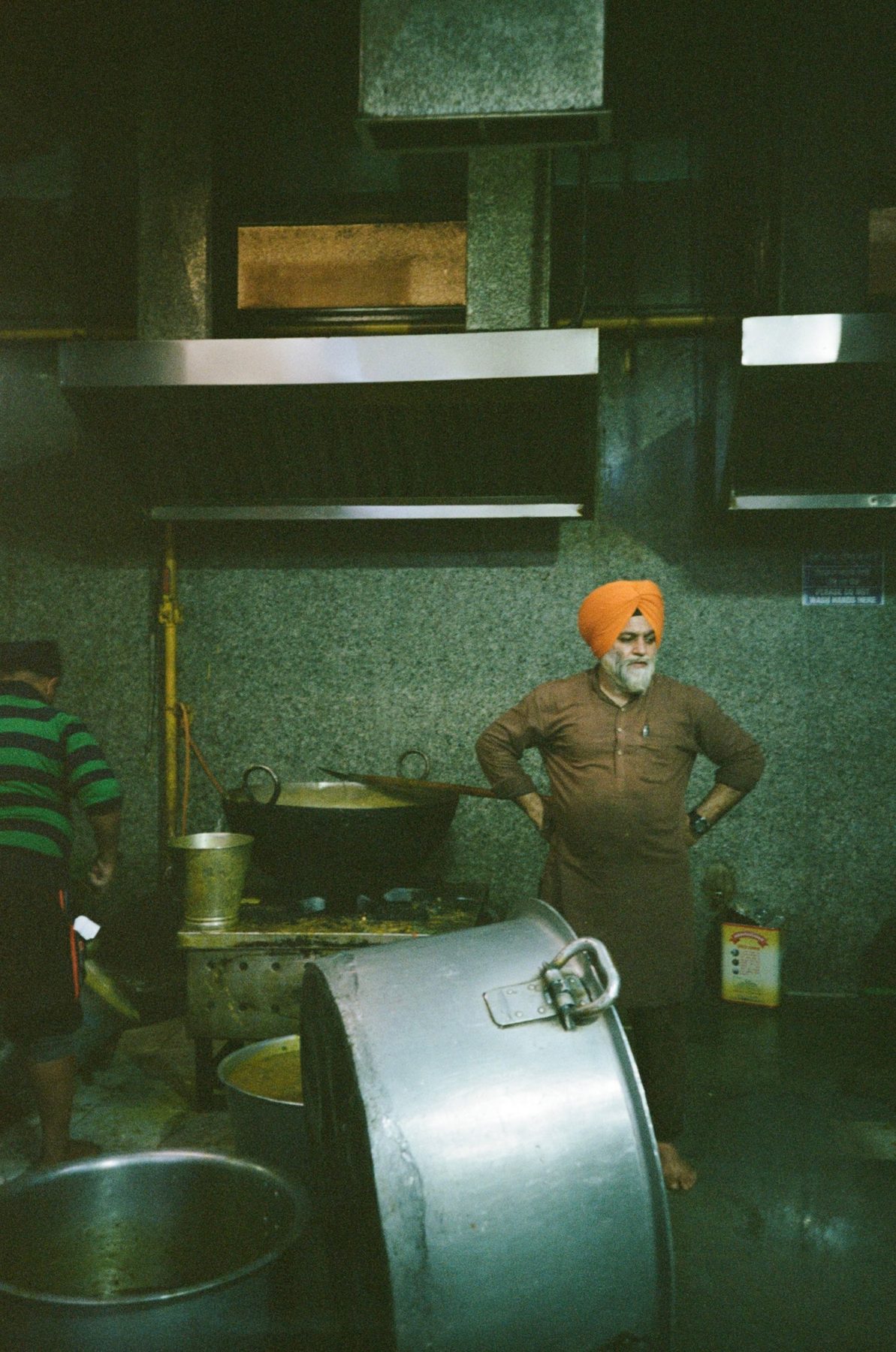 A Sikh cook in an orange turban, hands on hips, in a langar kitchen