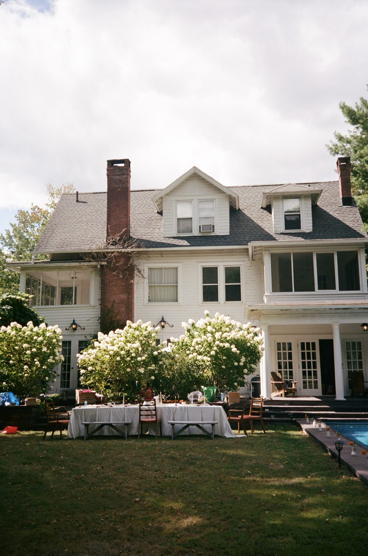 A white clapboard house with brick chimneys, hydrangeas and a set table on the lawn