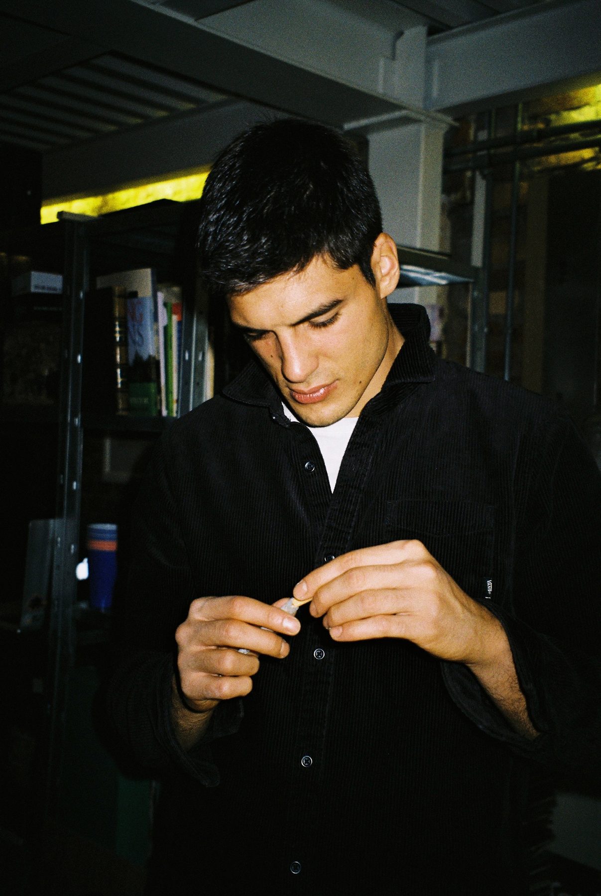 A young man in a black corduroy shirt looking down, lit by flash