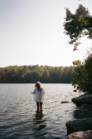 A woman in a white shirt wading into a lake ringed by trees