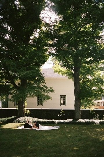 Two friends on a blanket in the shade of tall trees beside a white house