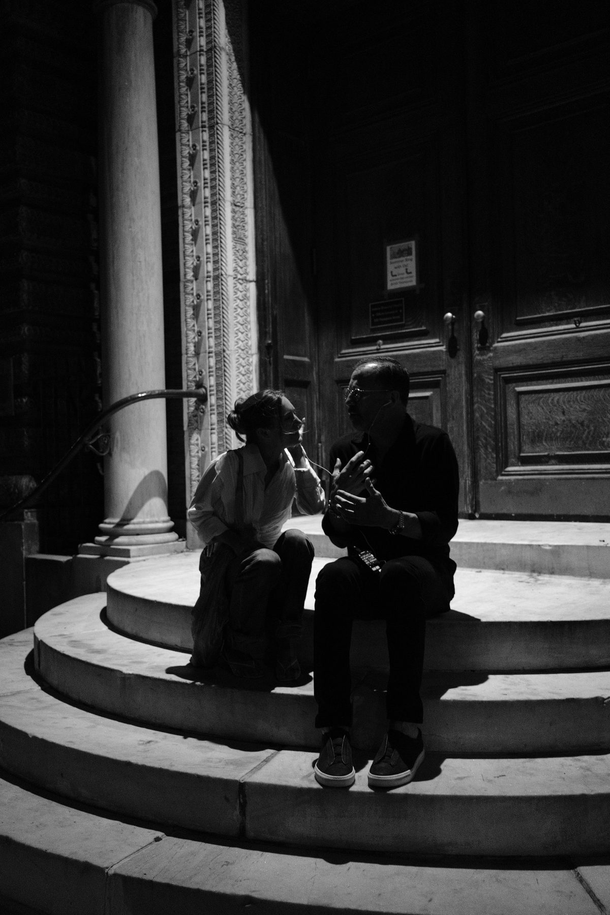 Two people talking on curved church steps in a hard wedge of light