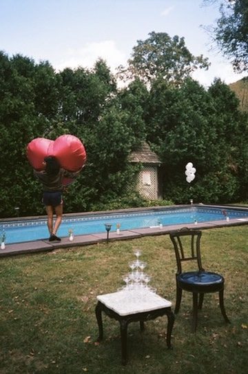 A woman carrying a giant red heart balloon past a pool, a champagne tower on the lawn
