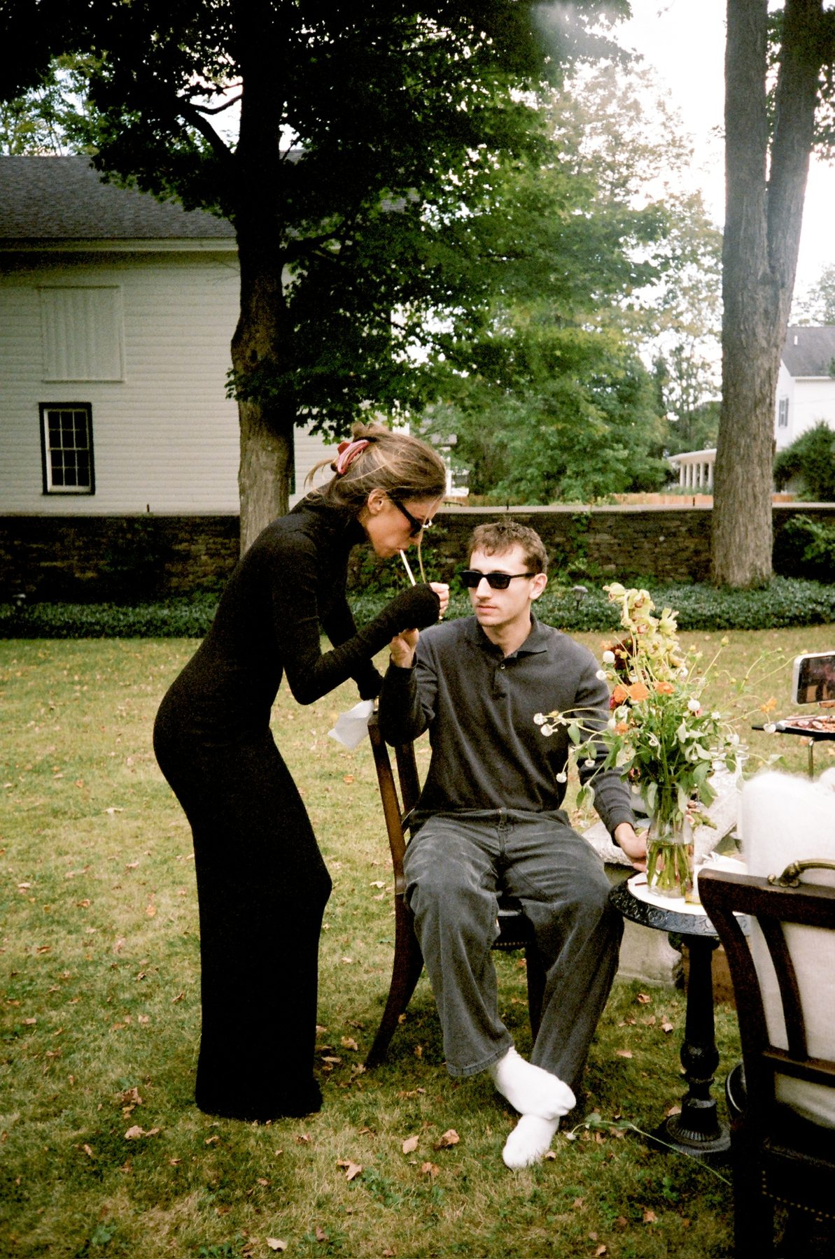 A woman in a black dress lighting a cigarette for a man in sunglasses on a garden chair