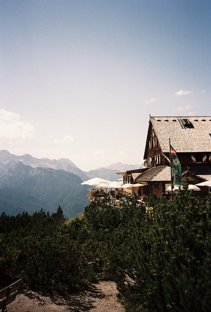 A shingled mountain hut with a busy terrace and white umbrellas above hazy peaks