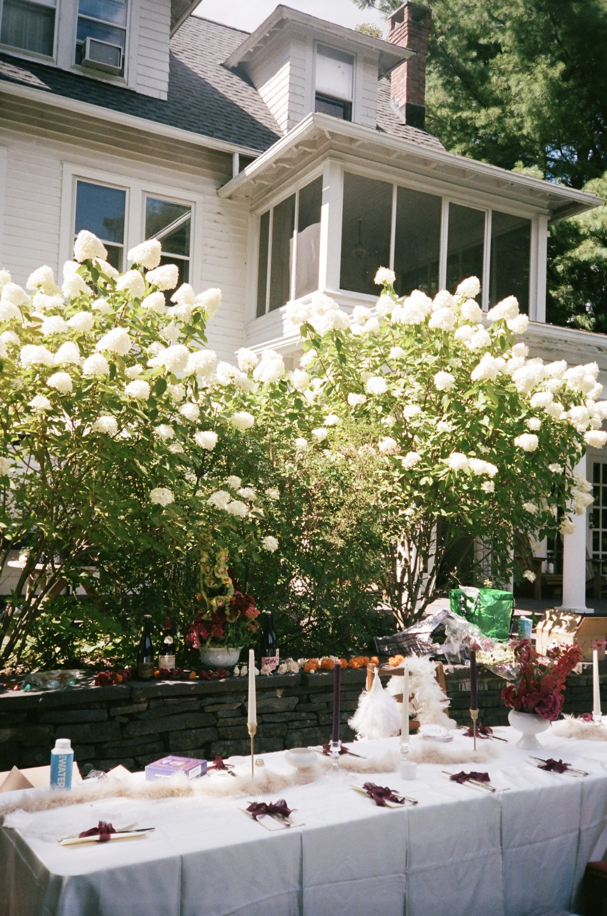 A long table set with candles and feathers before white hydrangeas and a clapboard house