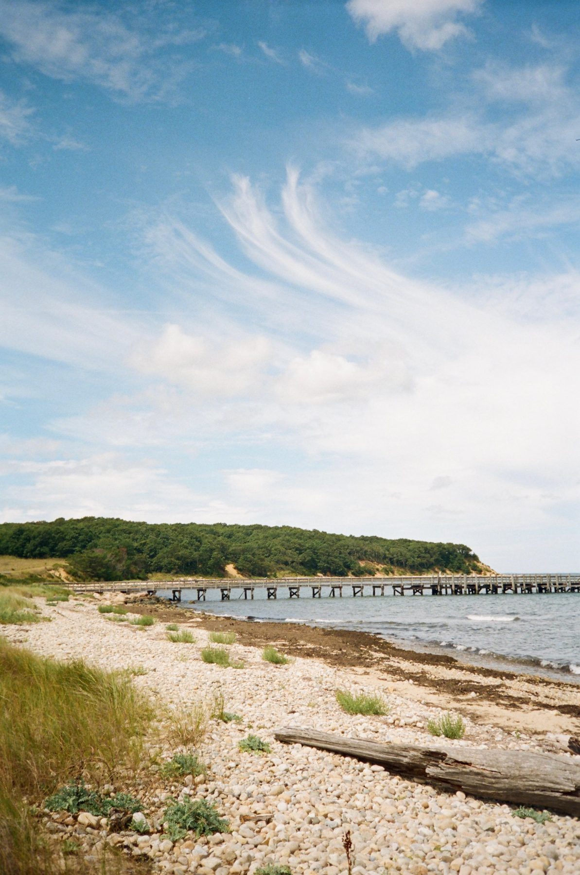 A long wooden pier off a pebble beach under sweeping white clouds