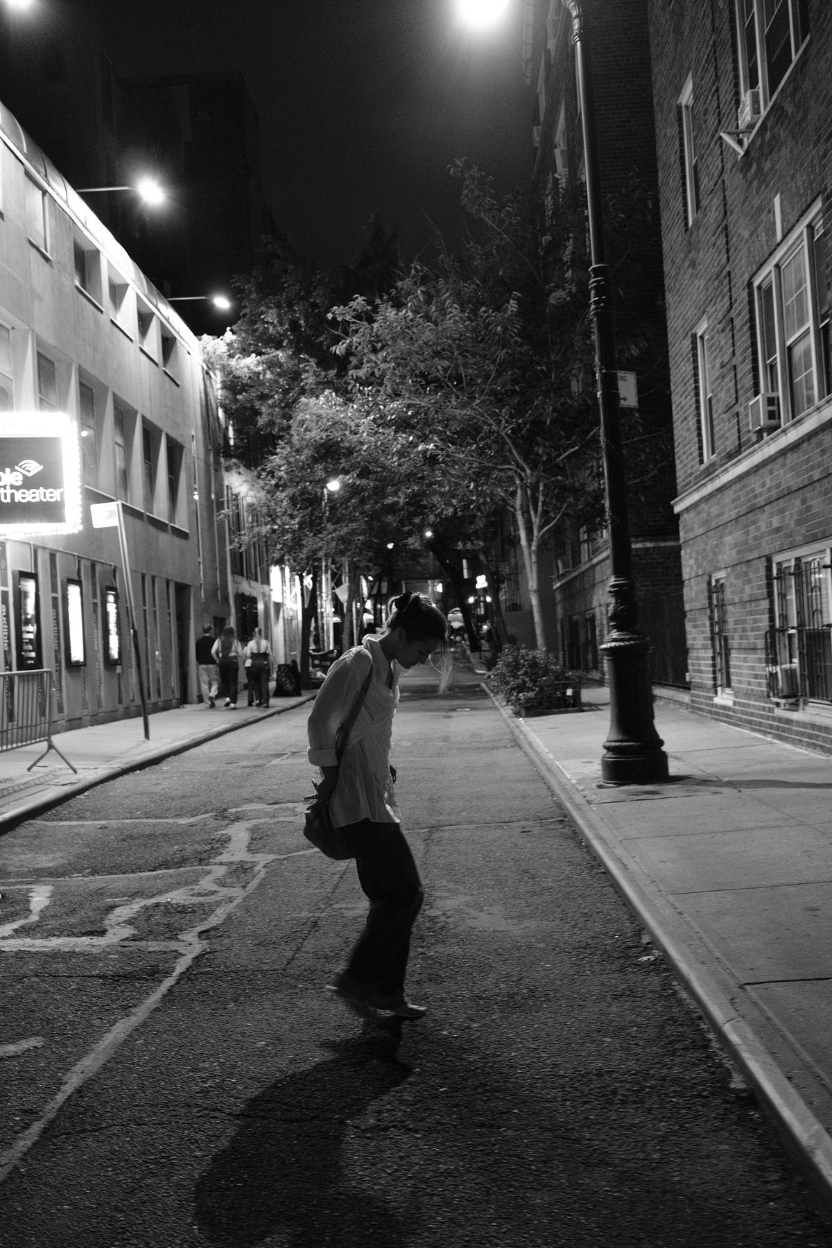 A woman looking down as she crosses an empty street beneath trees and streetlights