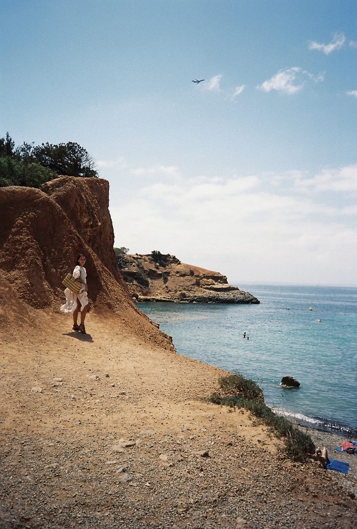 A woman in a white dress with a straw bag glancing back beside a red cliff, a plane overhead
