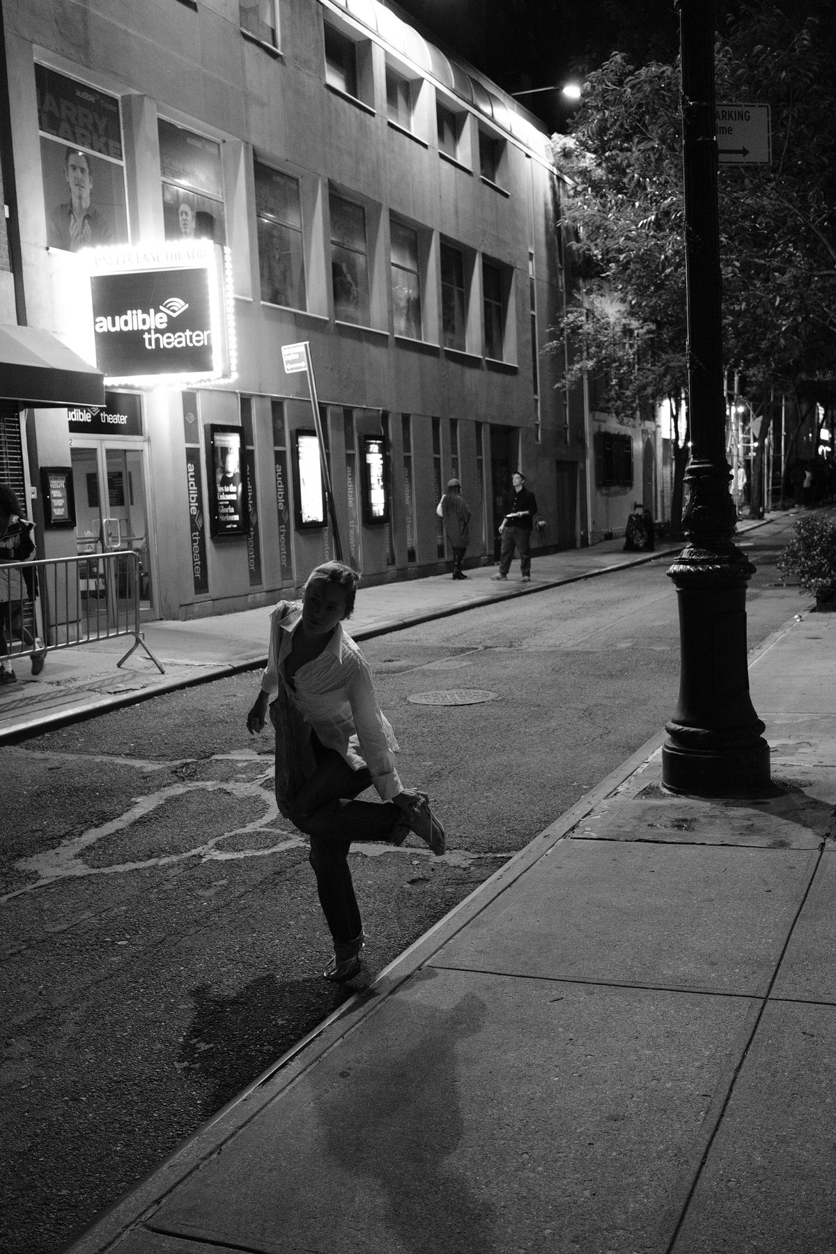 A woman kicking up one foot at the curb under a glowing theater marquee