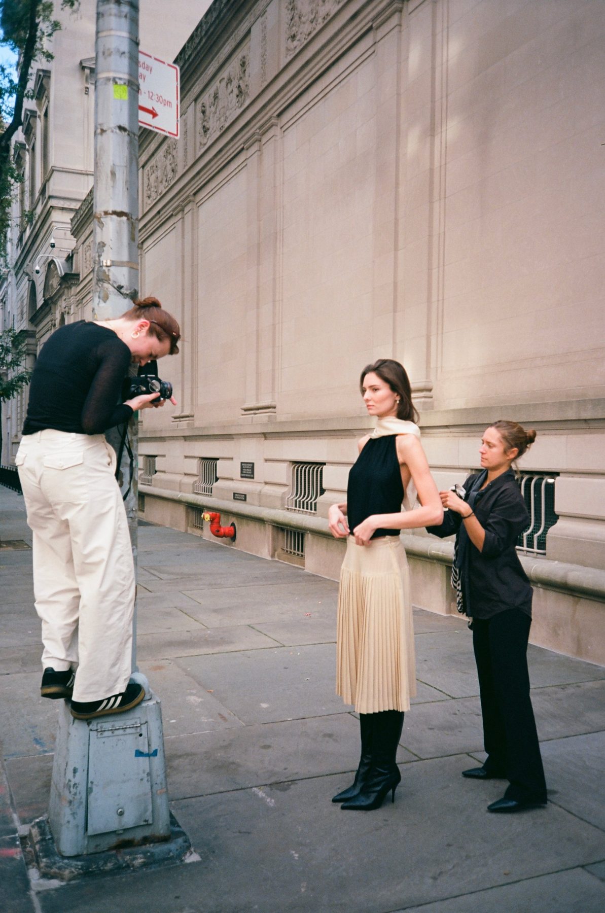 A photographer standing on a lamppost base shooting a model while a stylist adjusts her scarf