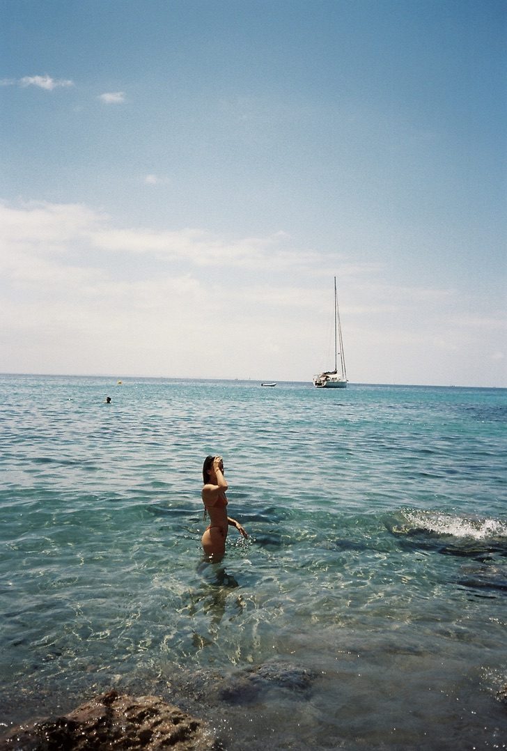 A woman standing waist-deep in clear water, a sailboat anchored on the horizon