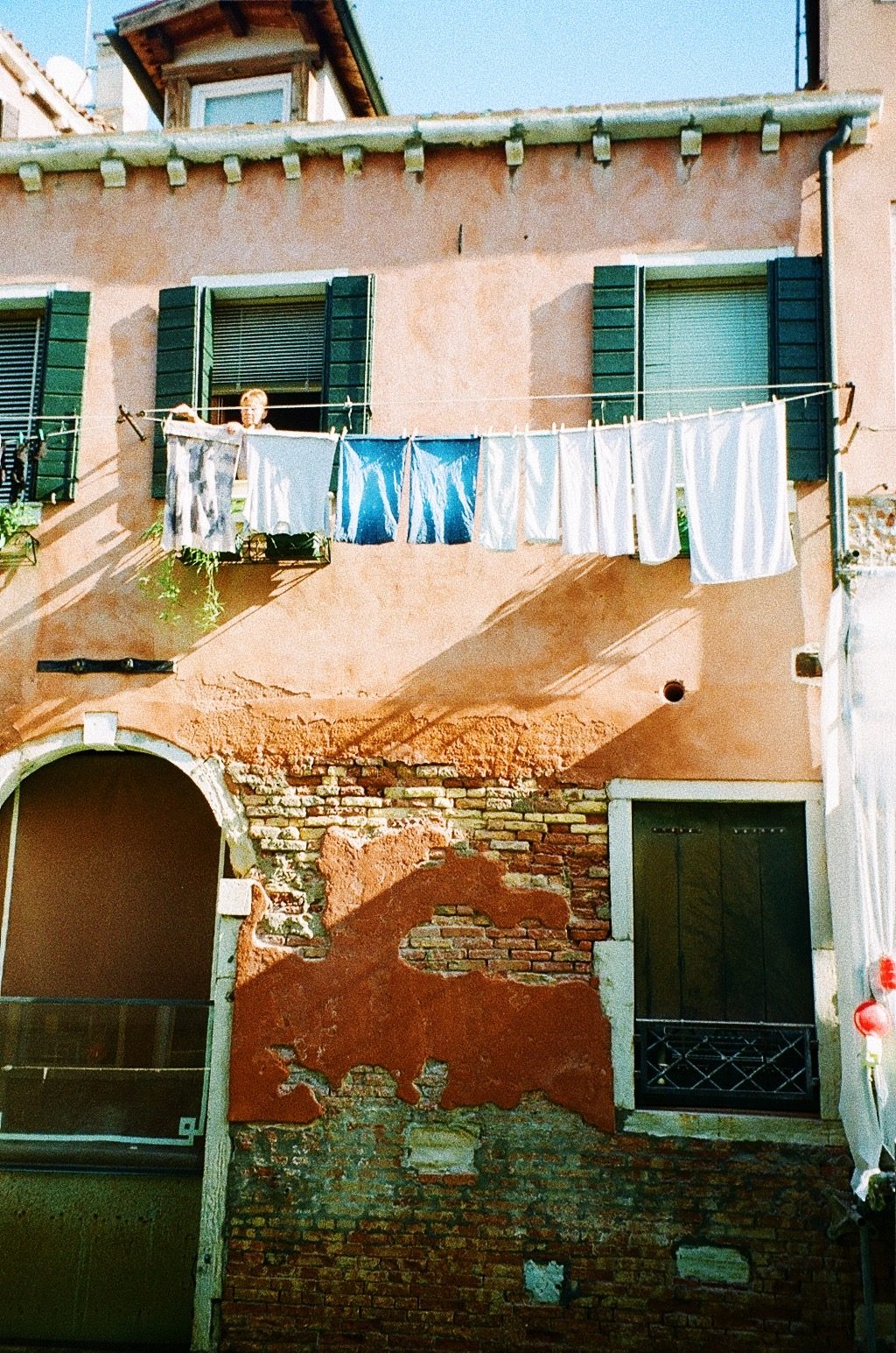 Laundry strung across a weathered terracotta facade with green shutters, a woman at the window