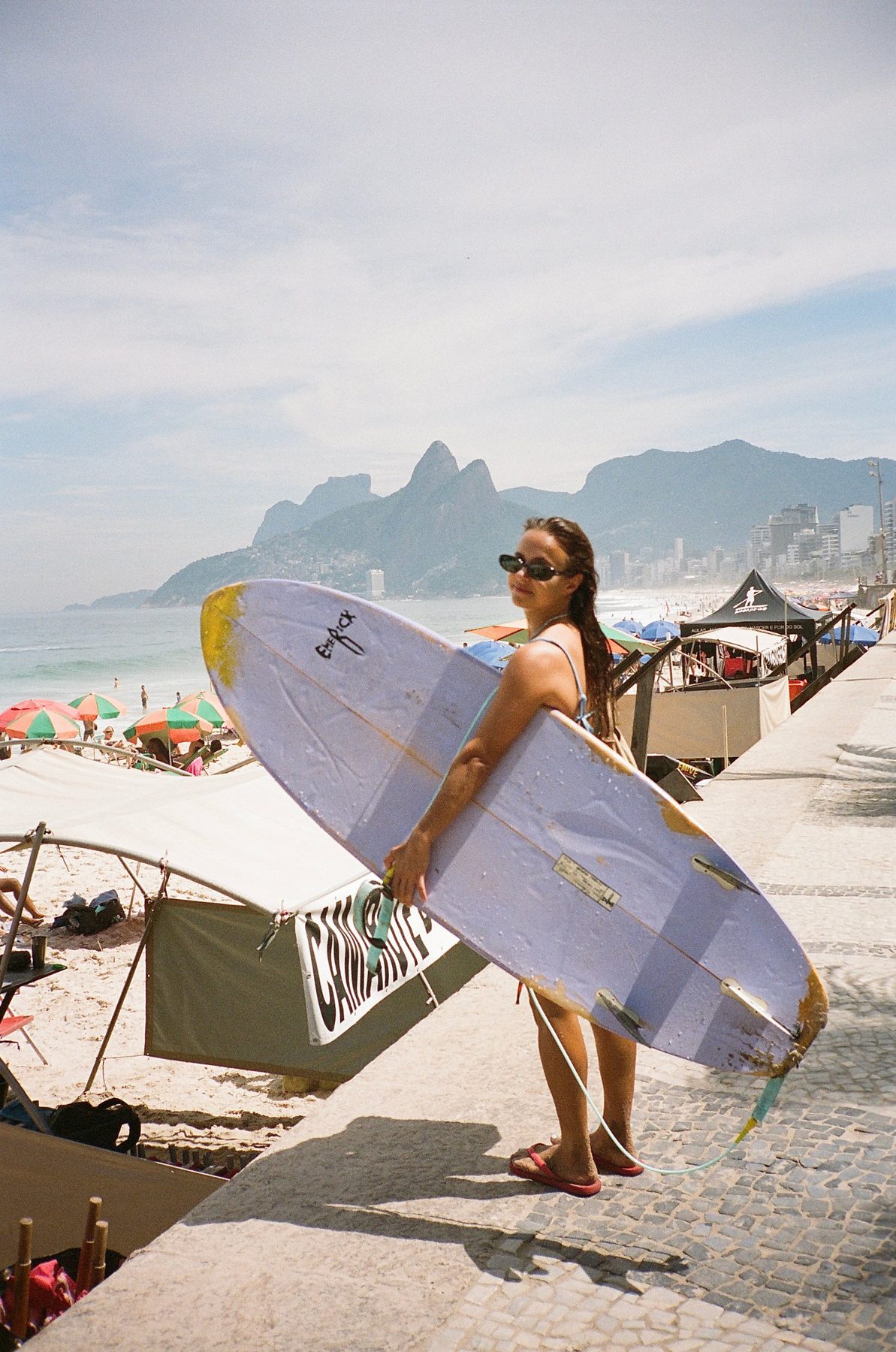 A woman in sunglasses holding a lilac surfboard on the Ipanema boardwalk, Dois Irmãos behind