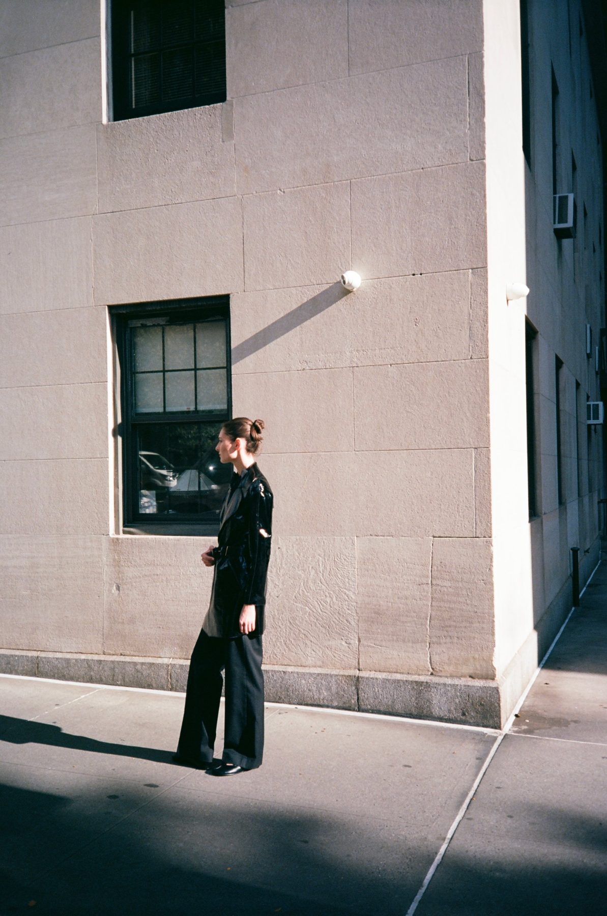 A woman in a black patent coat standing in hard light against a stone corner