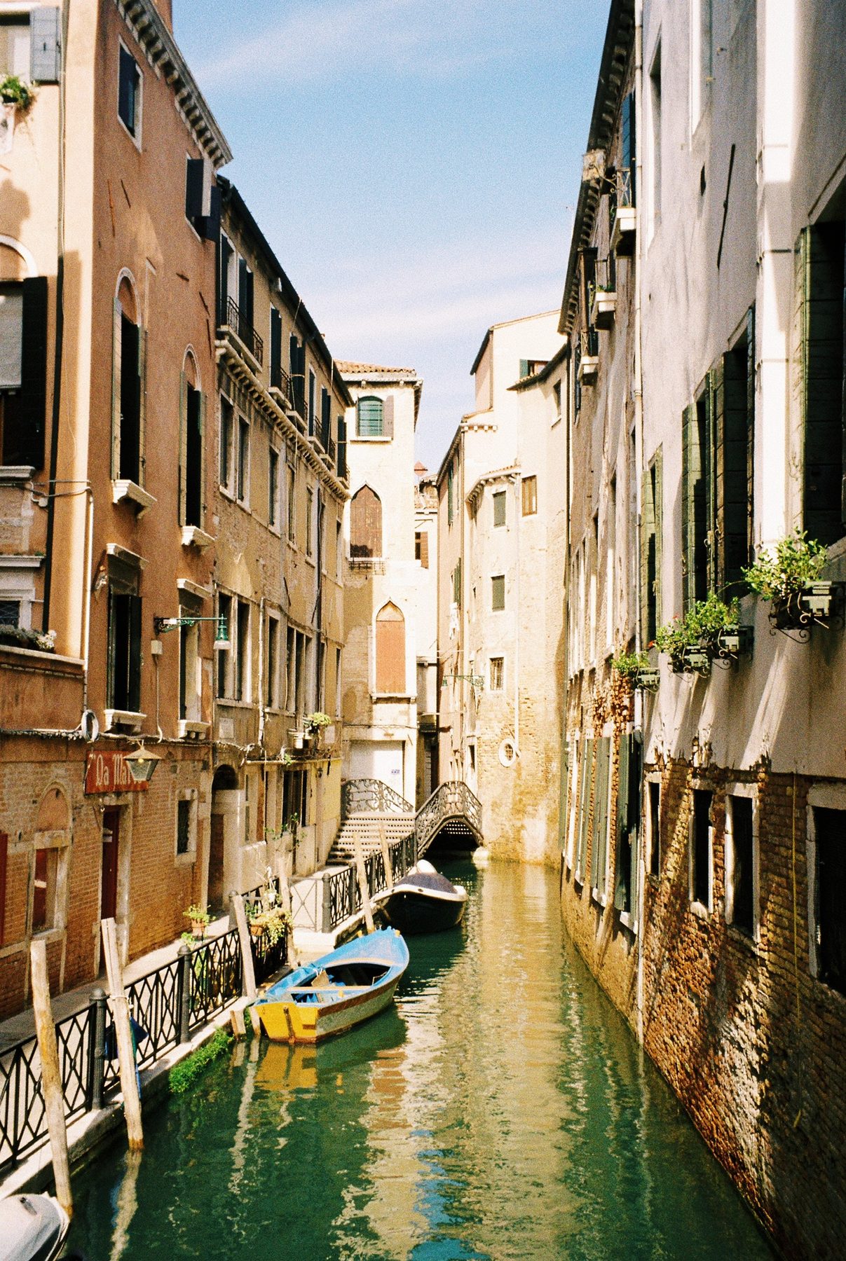 A yellow and blue rowboat in a narrow green canal below an iron bridge