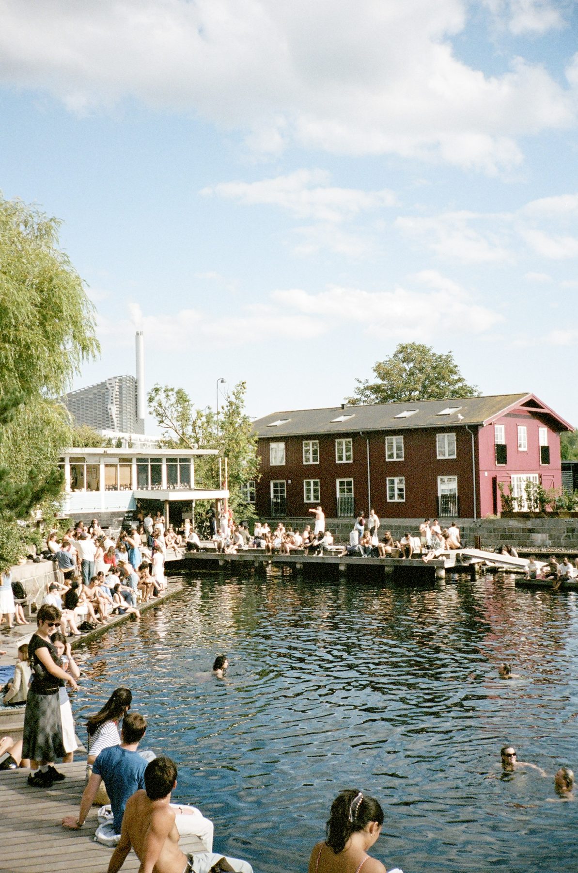Swimmers and sunbathers crowding a Copenhagen harbor bath beside a red wooden house