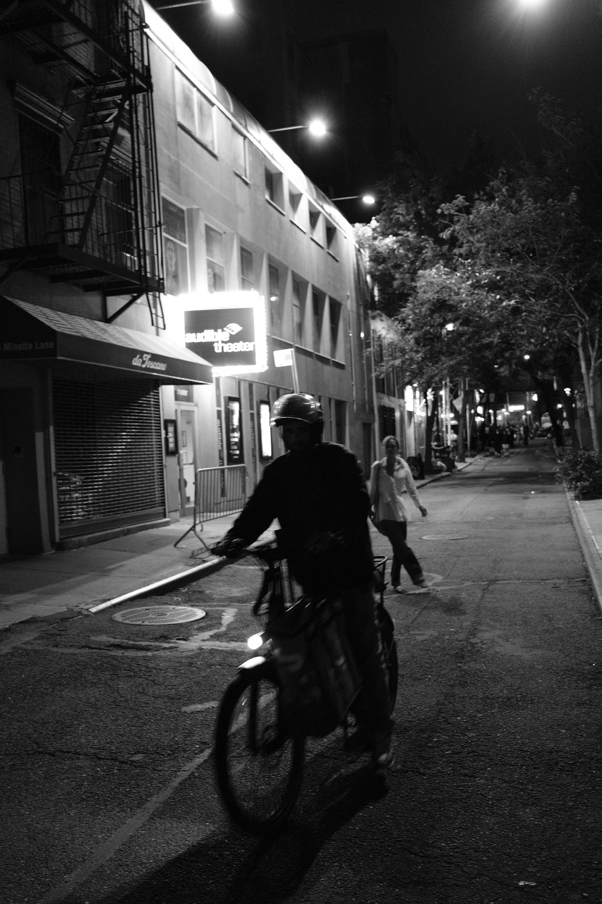 A delivery cyclist in silhouette passing the theater as a woman walks behind