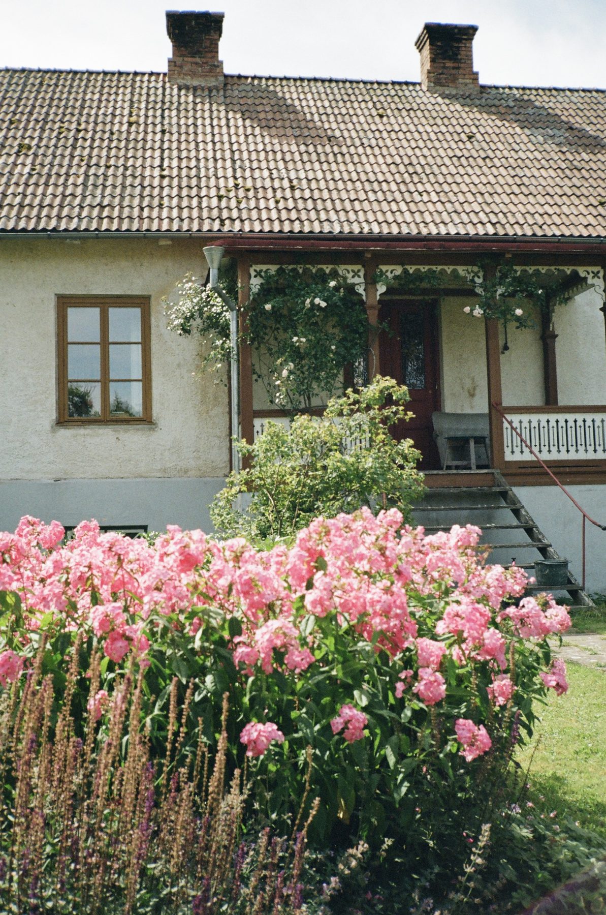 A cream farmhouse with a carved wooden porch behind a bank of pink phlox