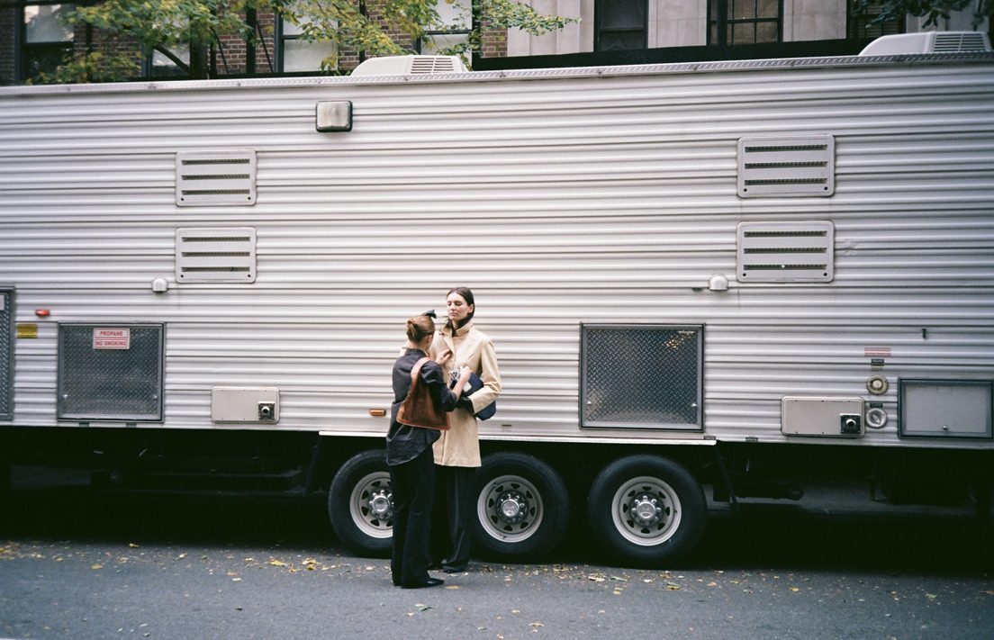 A stylist fixing a model’s coat beside a long silver production trailer