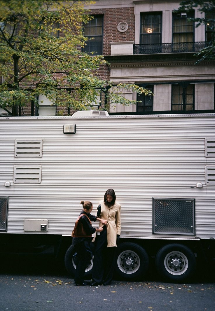 A stylist adjusting a model’s sleeve against the trailer under yellowing trees