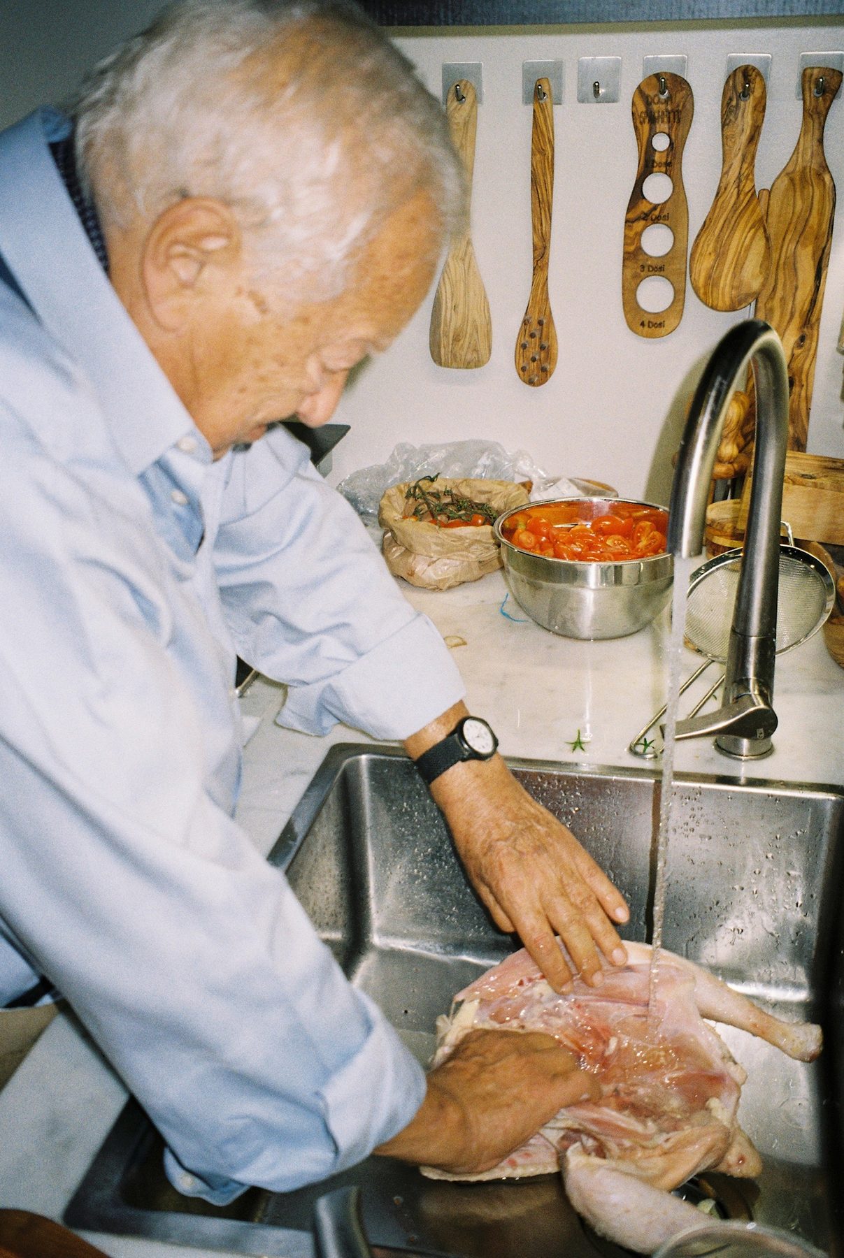 An older man in a blue shirt rinsing a chicken at the sink, cherry tomatoes and olive-wood spoons behind