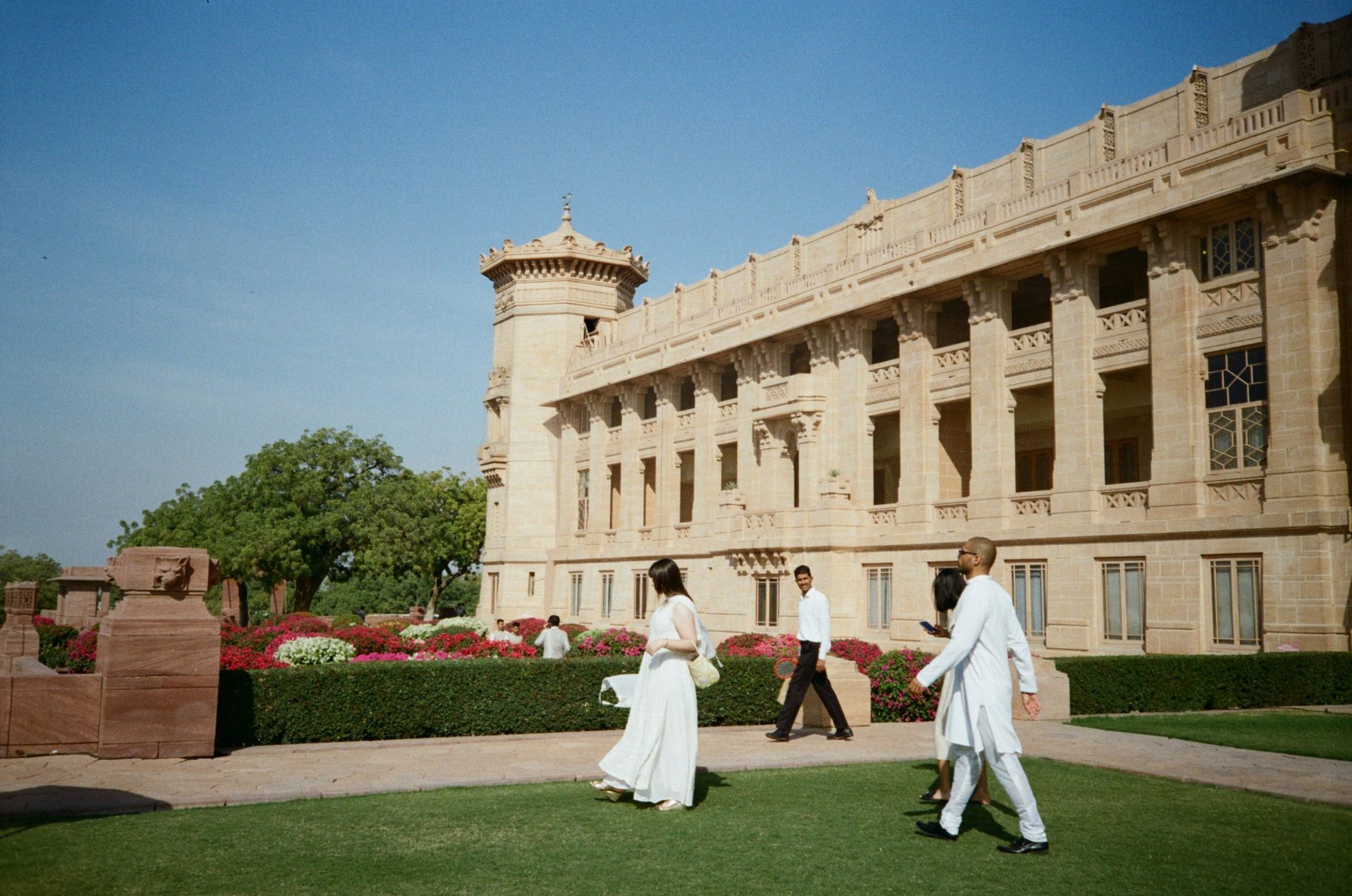 Guests in white walking across a lawn in front of a sandstone palace