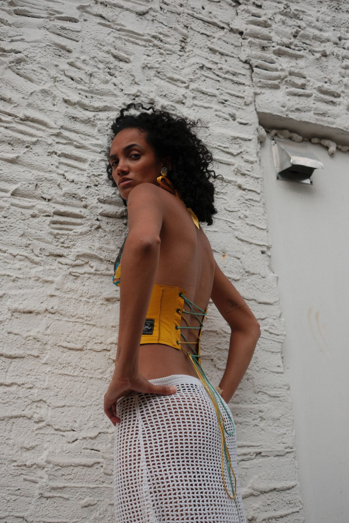 A woman glancing back over her shoulder against a textured white wall, corset laced in green and yellow