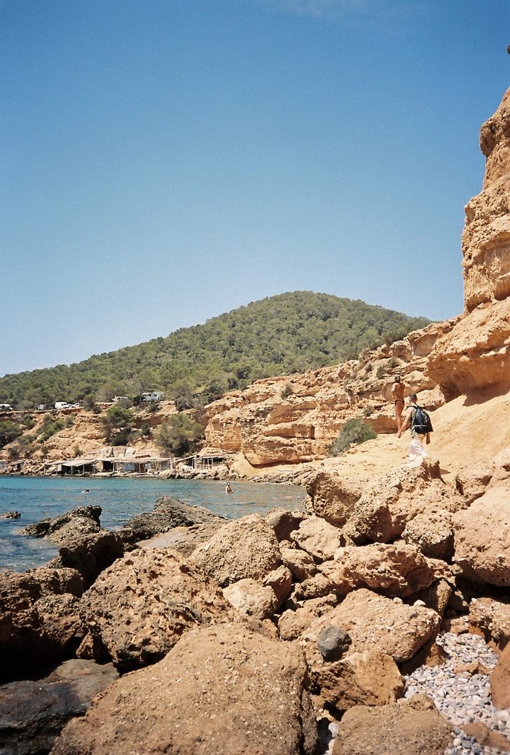 Two figures climbing sandstone rocks above a turquoise cove with fishermen's huts