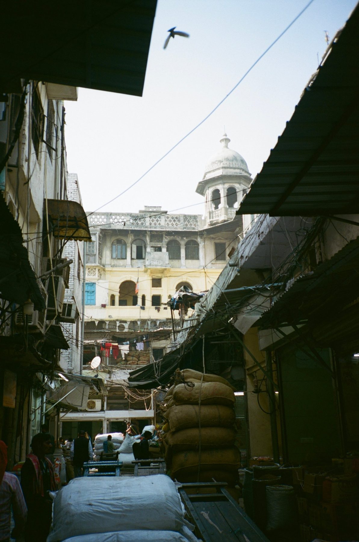 A market lane piled with sacks below a domed old haveli, a bird overhead