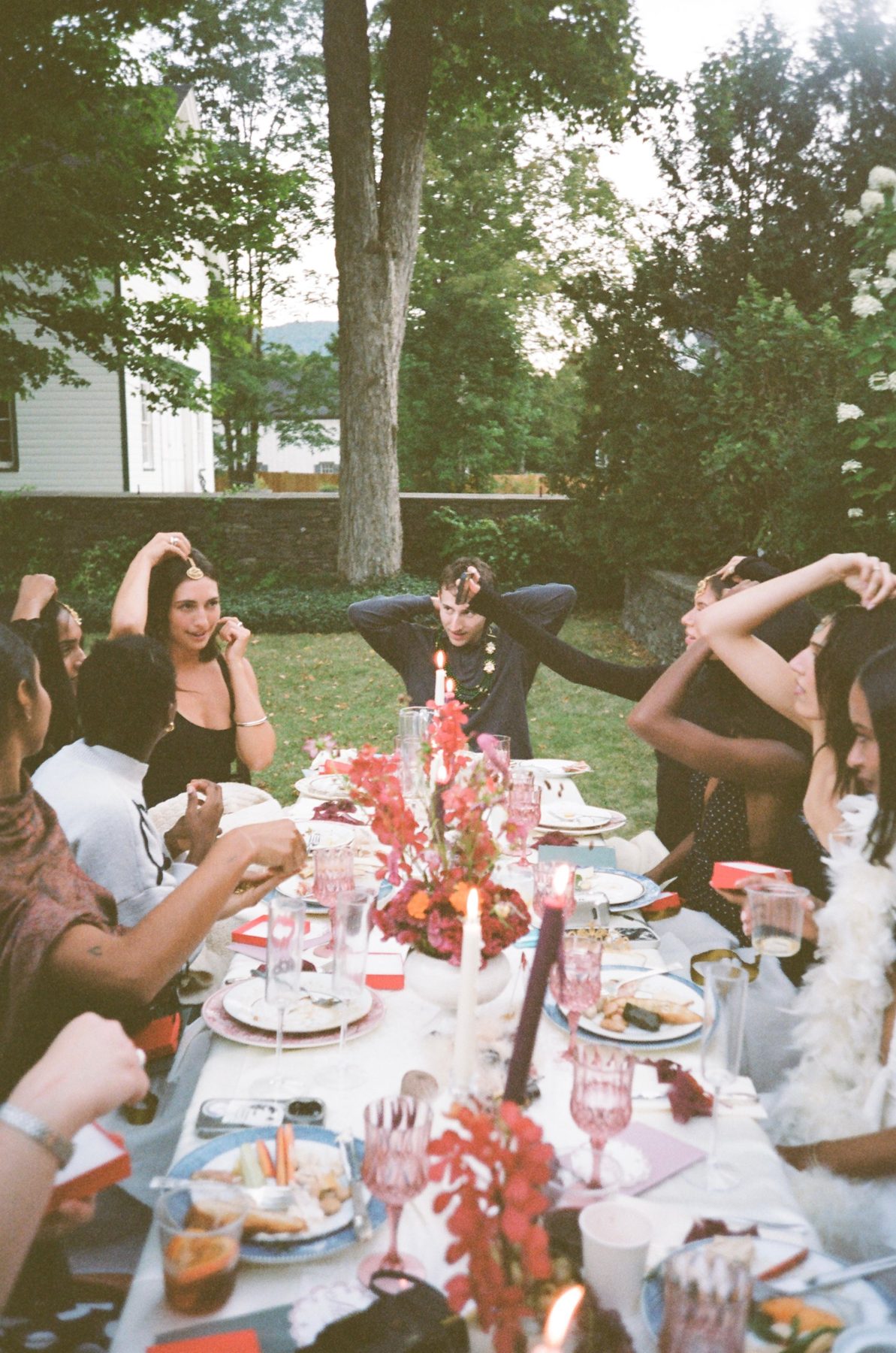 Friends at a long candlelit dinner table on a lawn, red flowers and pink glasses