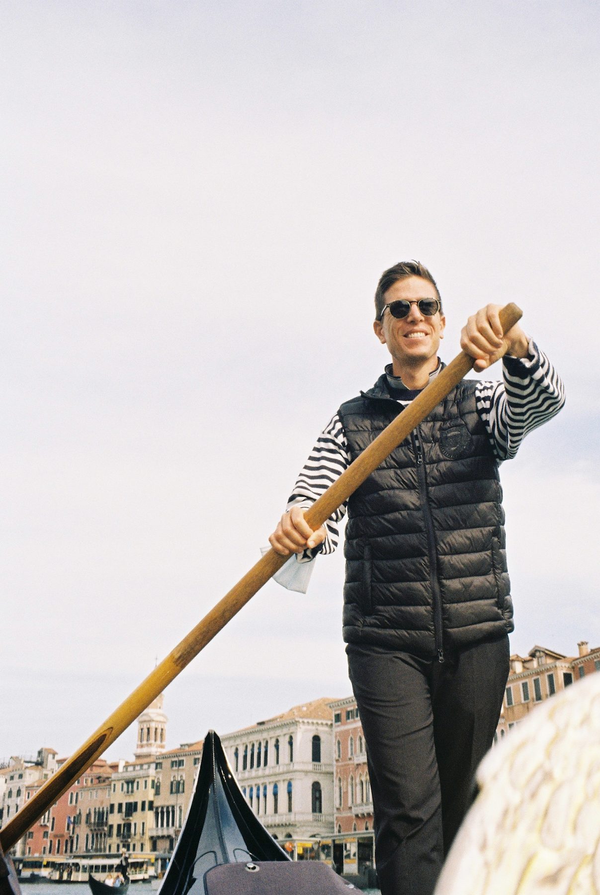 A smiling gondolier in a striped shirt and sunglasses rowing on the Grand Canal