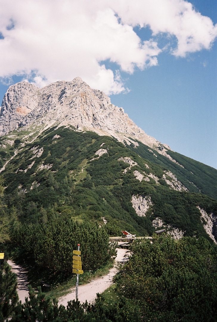 A yellow trail sign at a bend in a mountain road below a rocky summit
