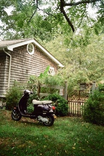 A purple Vespa parked on the grass beside a shingled cottage under leafy branches