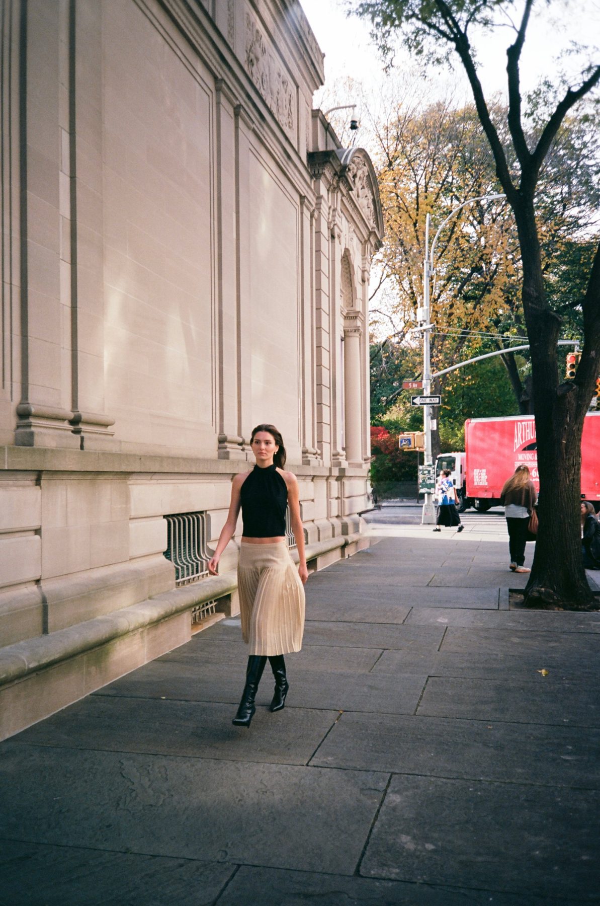 A model in a black halter and pleated skirt walking along a limestone wall on Fifth Avenue