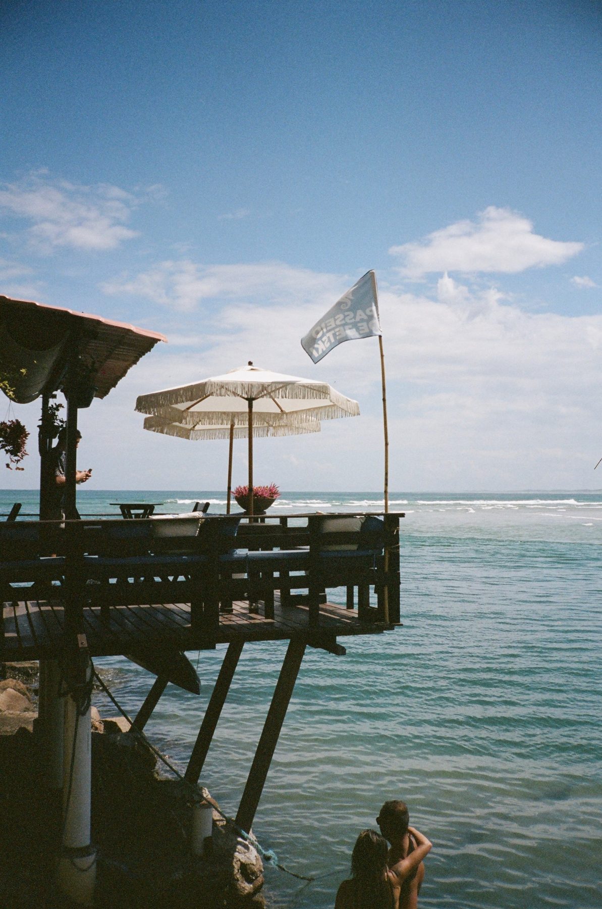 A wooden deck with fringed umbrellas over turquoise water, a couple below