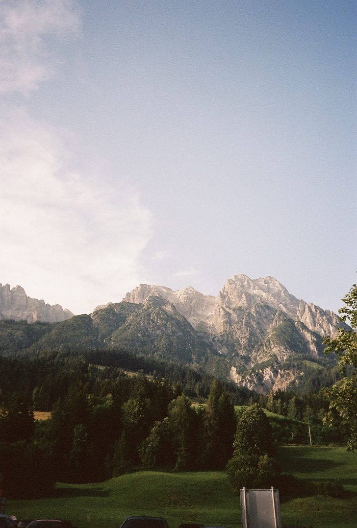 Soft grey peaks rising over a green meadow and forest under a pale sky