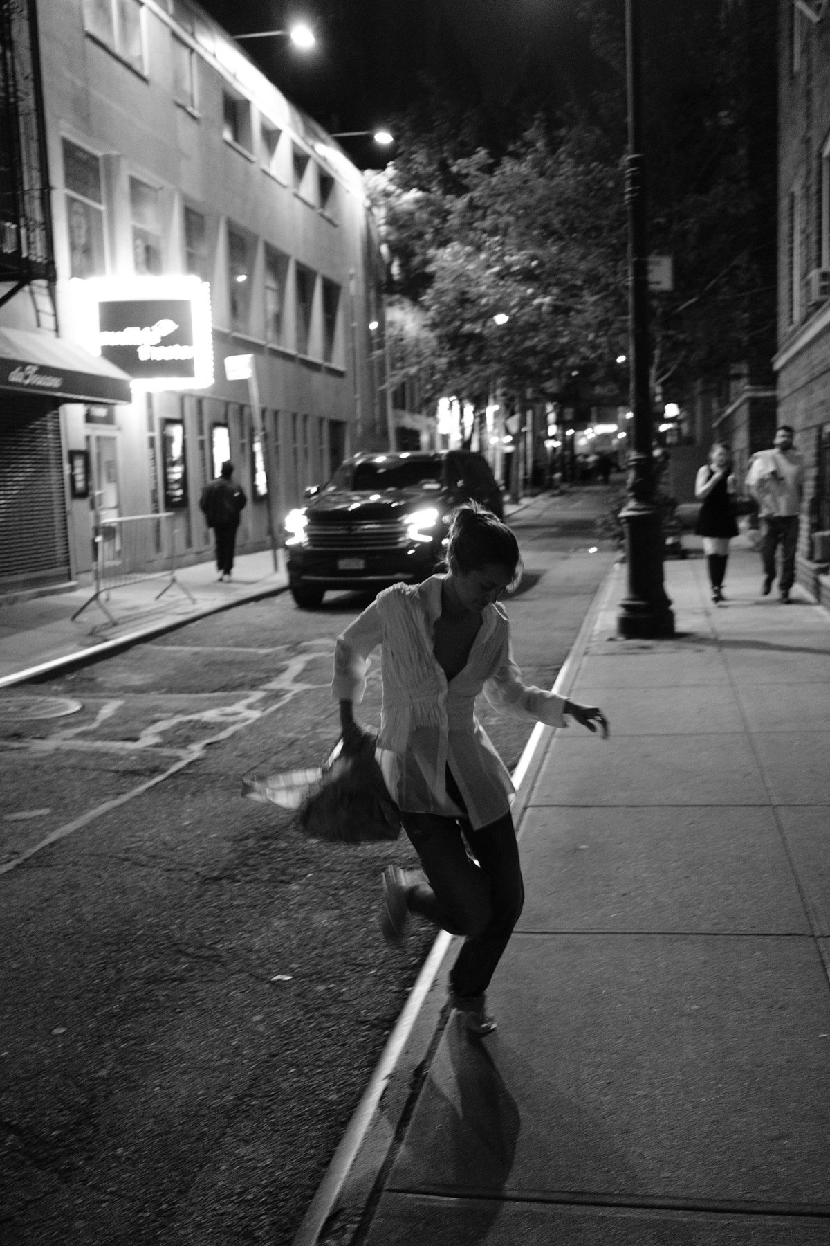 A woman in a white shirt dancing off the curb on a lamplit street outside a theater