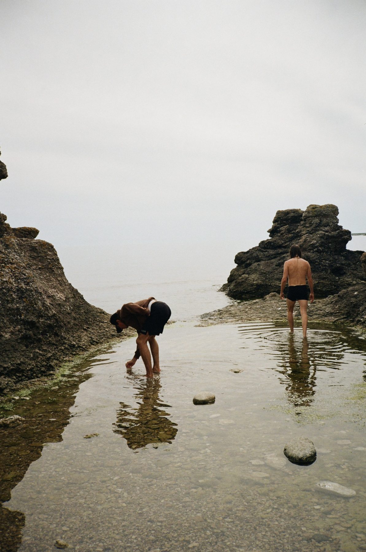 Two men wading in a still tide pool between dark rocks under a white sky
