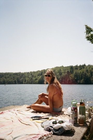 A woman in sunglasses on a towel at the edge of a lake on a bright day