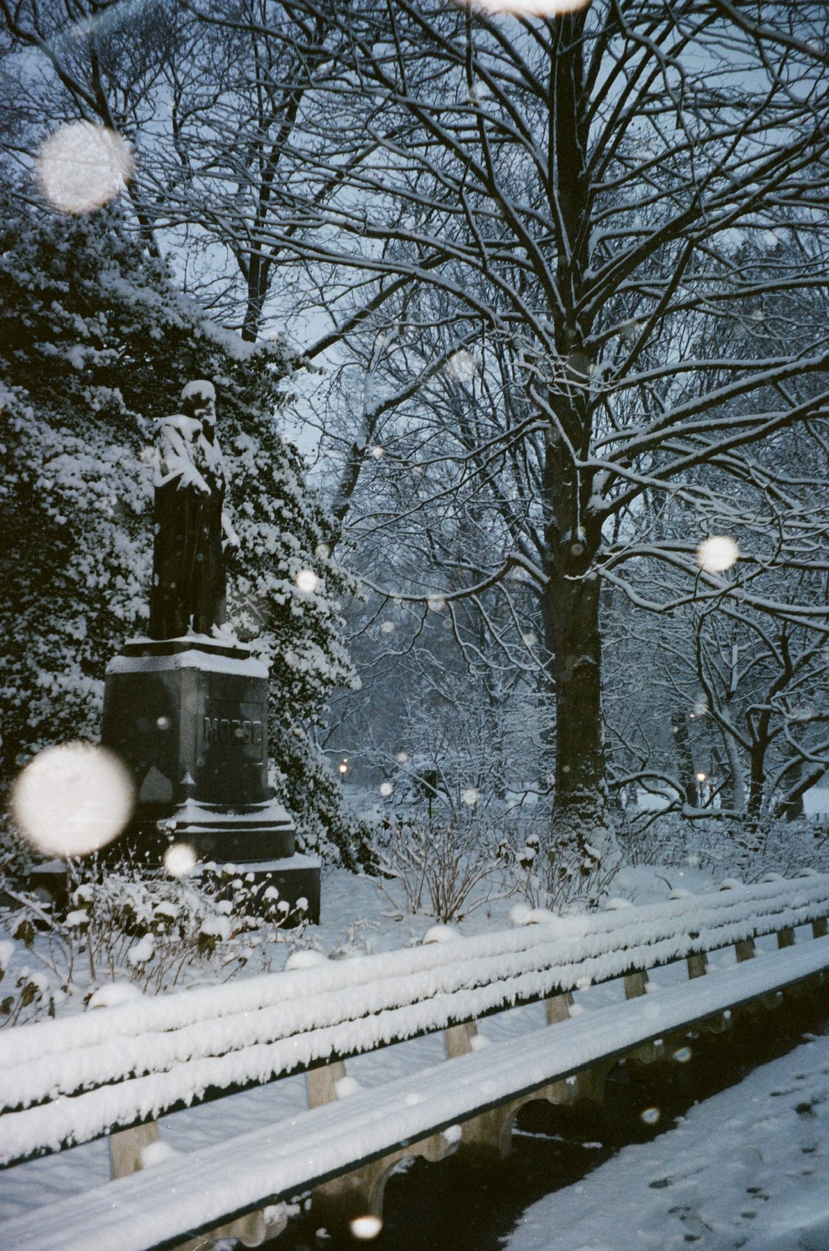 A snow-capped statue and long bench in the park at dusk, snowflakes in the air