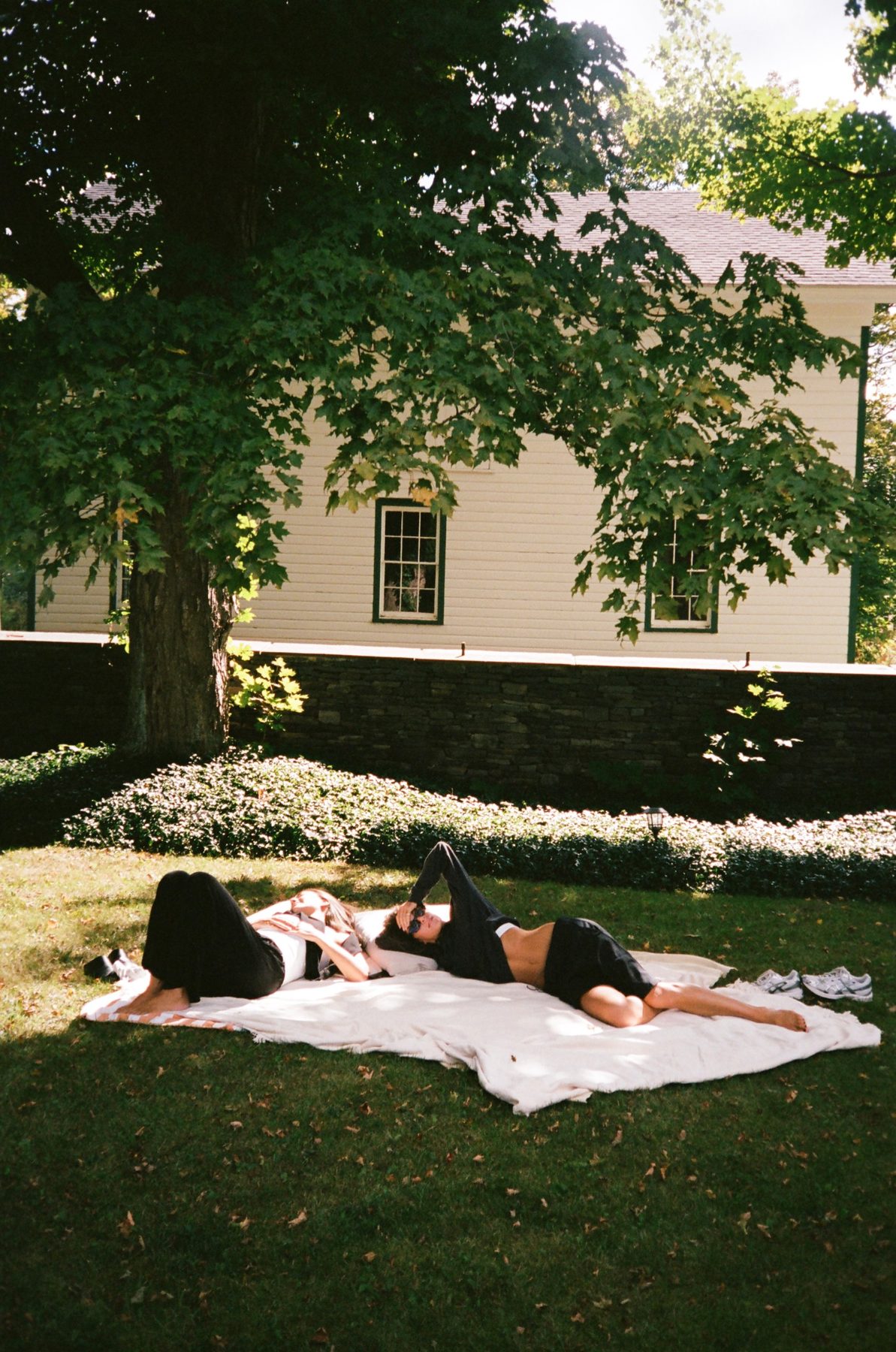 Two women lying on a blanket in the shade of a big tree by a white house