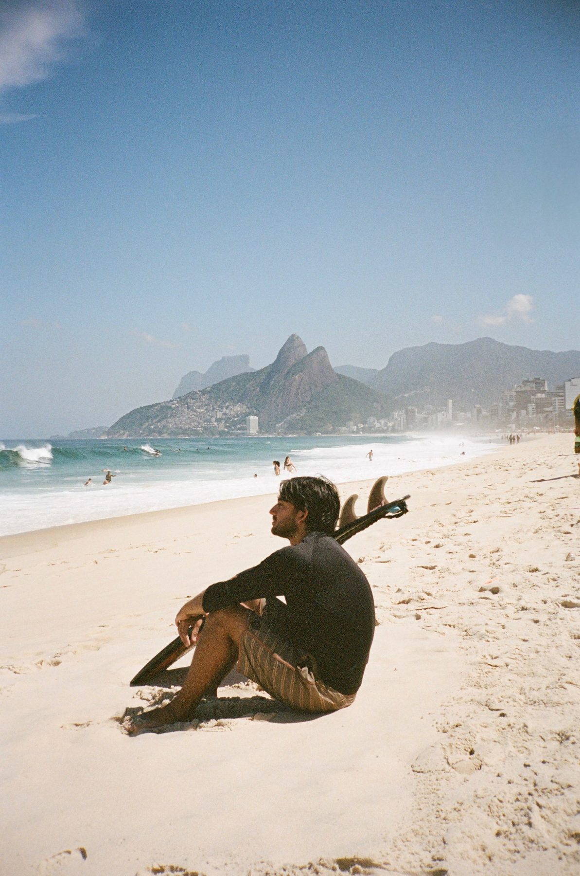 A surfer sitting in the sand beside his board, the beach curving away